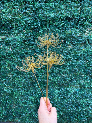 Three gold metal wire chrysanthemum stems held by a hand against a green leafy hedge background.
