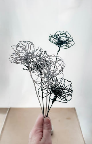A cluster of three silver wire flowers with intricate, openwork petals. The stems are long and slender, and the flowers are arranged in a bouquet. The background is a solid, dark blue. carnation January birth flower preserved florals Mother’s Day gifts gift for mom silver and black wire flowers delicate pretty decor wedding floral preservation forver flowers wire flower stem carnations curved leaf handmade small business steel aluminum petals dried florals whimsical decor