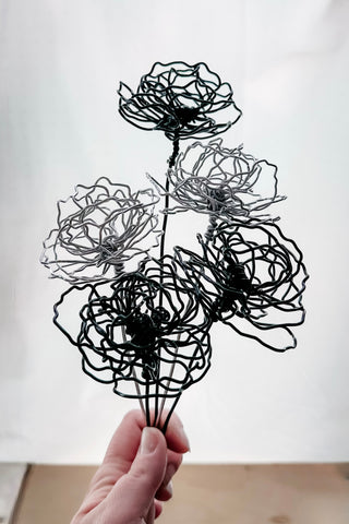 A cluster of three silver wire flowers with intricate, openwork petals. The stems are long and slender, and the flowers are arranged in a bouquet. The background is a solid, dark blue. carnation January birth flower preserved florals Mother’s Day gifts gift for mom silver and black wire flowers delicate pretty decor wedding floral preservation forver flowers wire flower stem carnations curved leaf handmade small business steel aluminum petals dried florals whimsical decor